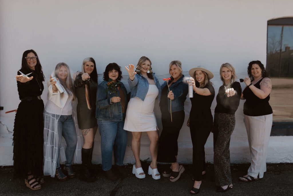 - Ibi David Salon in Westlake, OH Nine women stand in a row outdoors, facing the camera and extending their hands forward, some holding flowers or objects, against a plain white wall background. - Ibi David Salon in Westlake, OH