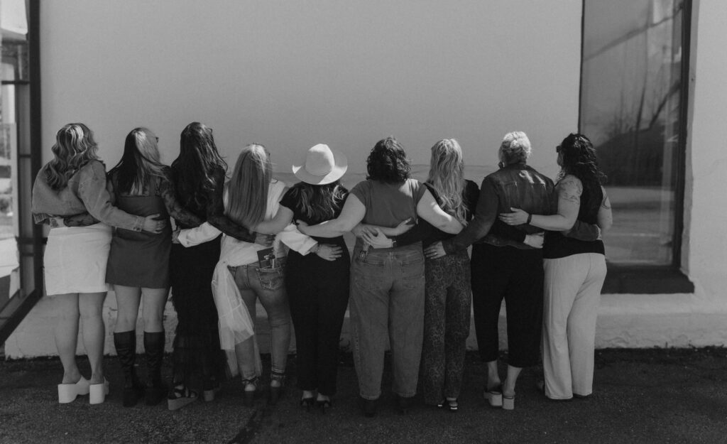 - Ibi David Salon in Westlake, OH Nine women stand side by side facing away from the camera, arms around each other, in front of a plain wall in an outdoor setting. - Ibi David Salon in Westlake, OH